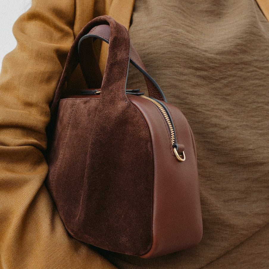 Model holding STOW London's Mini Bowling in Chocolate suede leather featuring a pleated front panel and gold-coated stainless steel hardware