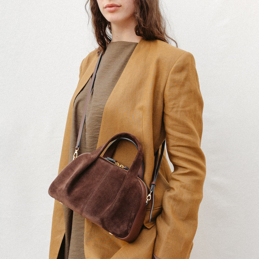 Model wearing STOW London's Mini Bowling in Chocolate suede leather crossbody featuring a pleated front panel and gold-coated stainless steel hardware