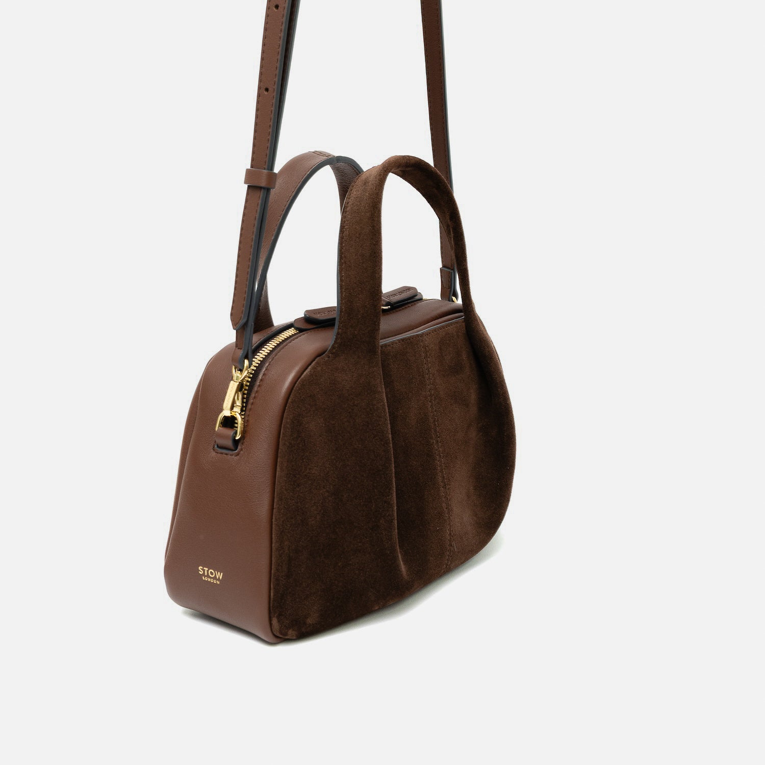 Off-axis view of STOW London's Mini Bowling bag in Chocolate suede and smooth leather featuring a pleated front panel, detachable crossbody strap and gold-coated stainless steel hardware on a light grey background