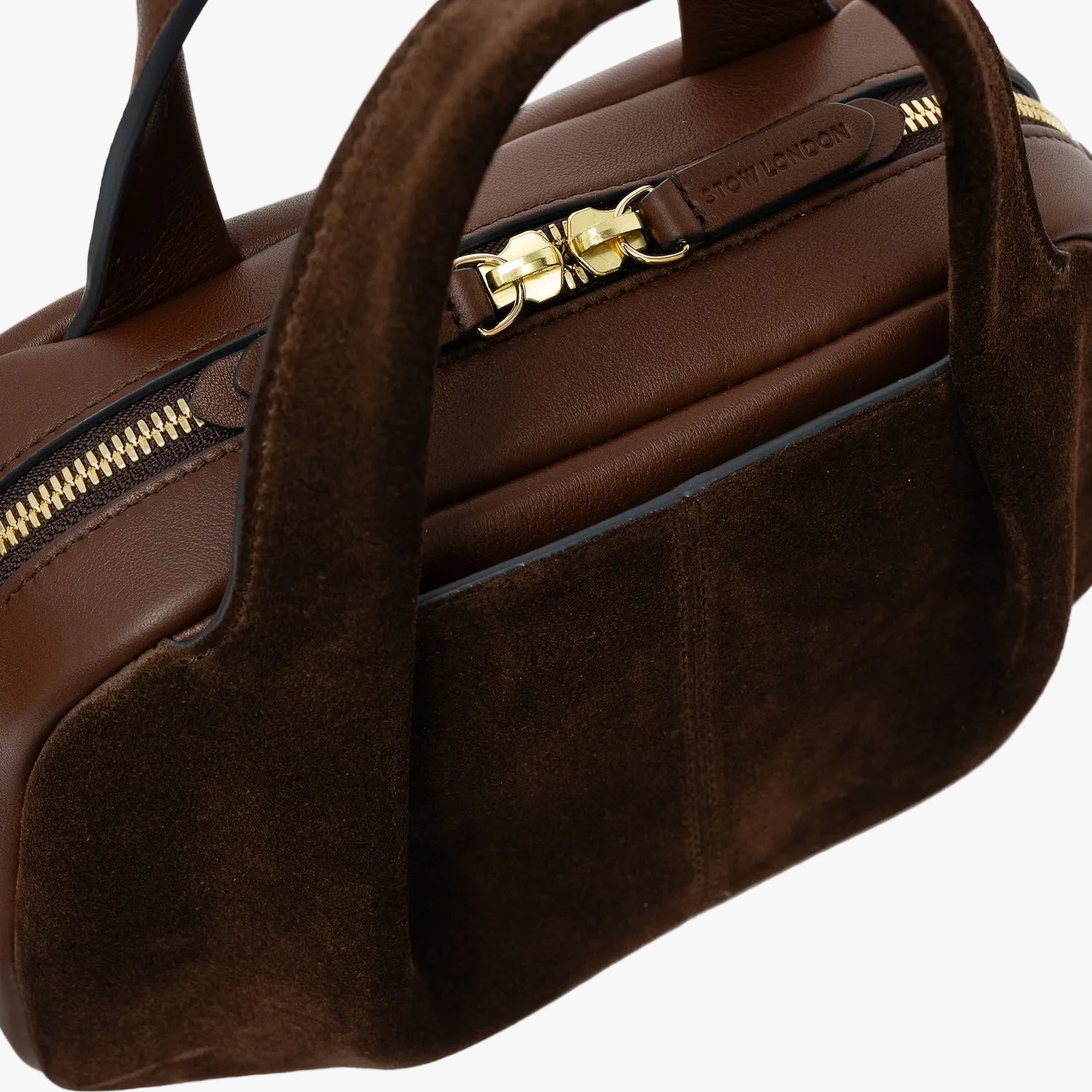 Close up of STOW London's Mini Bowling bag in Chocolate suede and smooth leather featuring a pleated front panel with slip pocket in-between, zip closure and STOW London embossed pull tabs on a light grey background