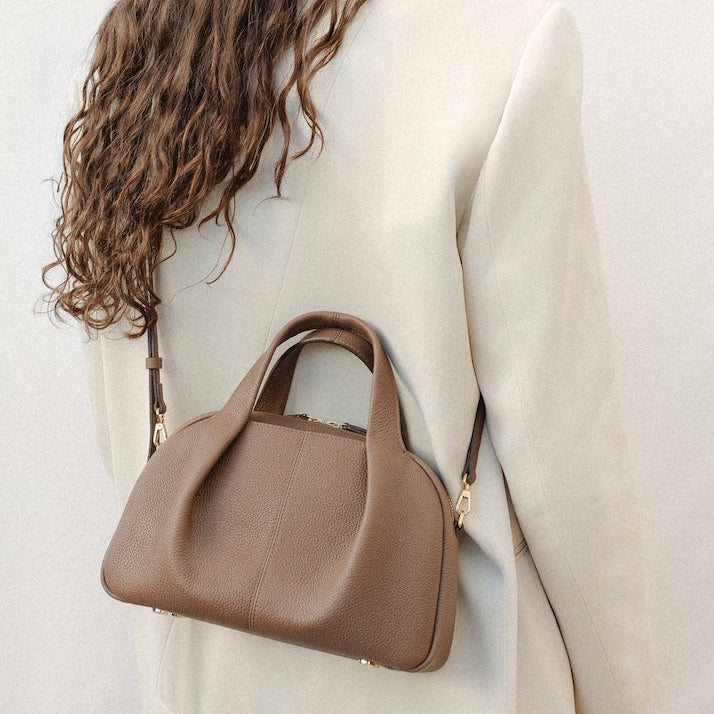 Model wearing STOW London's Mini Bowling bag in Camel pebbled leather crossbody featuring a pleated front panel, detachable crossbody strap and gold-coated stainless steel hardware