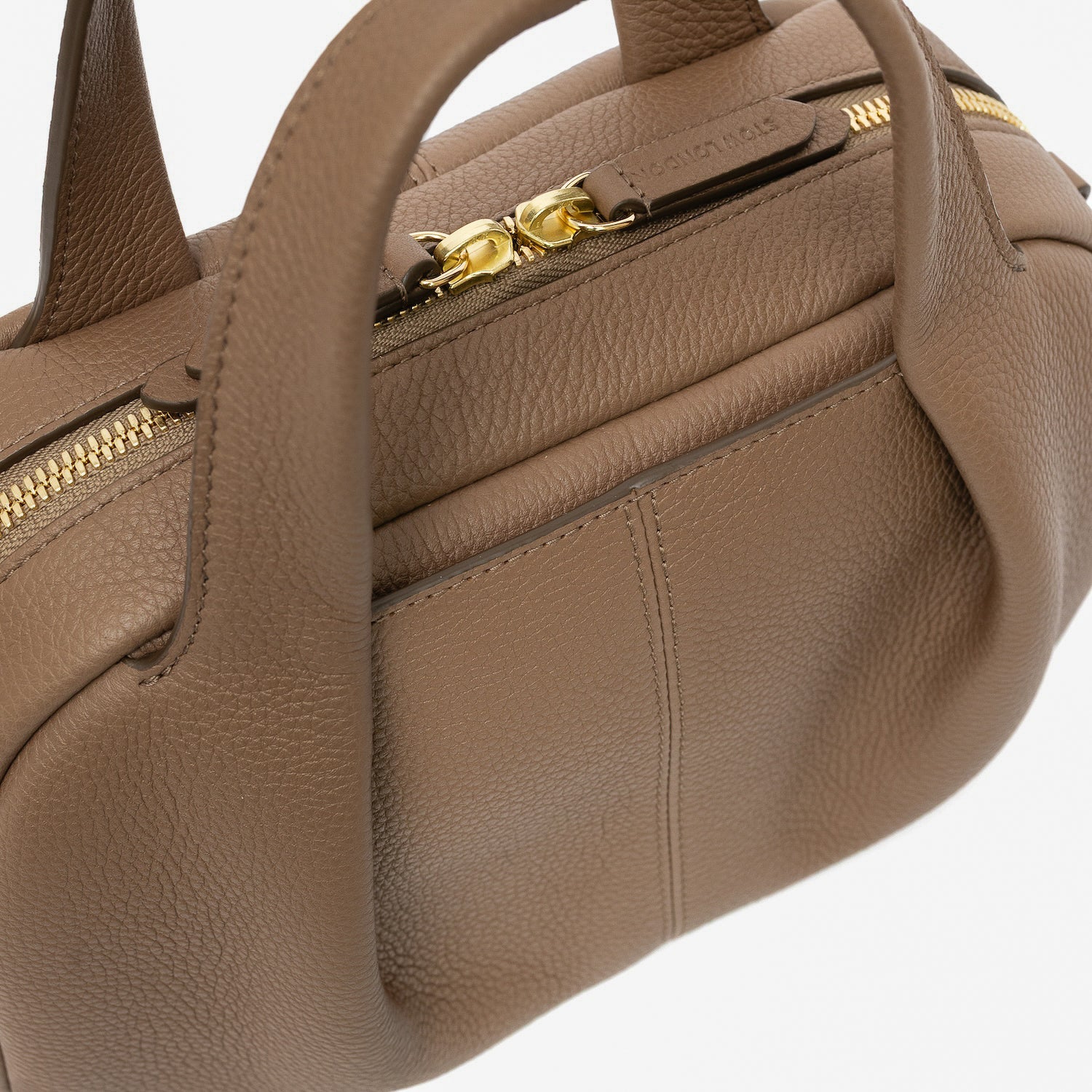 Close up of STOW London's Mini Bowling bag in Mocha pebbled leather featuring the pleated front panel with external pocket in-between, gold-coated stainless steel hardware, zip closure and STOW London embossed pull tabs on a light grey background