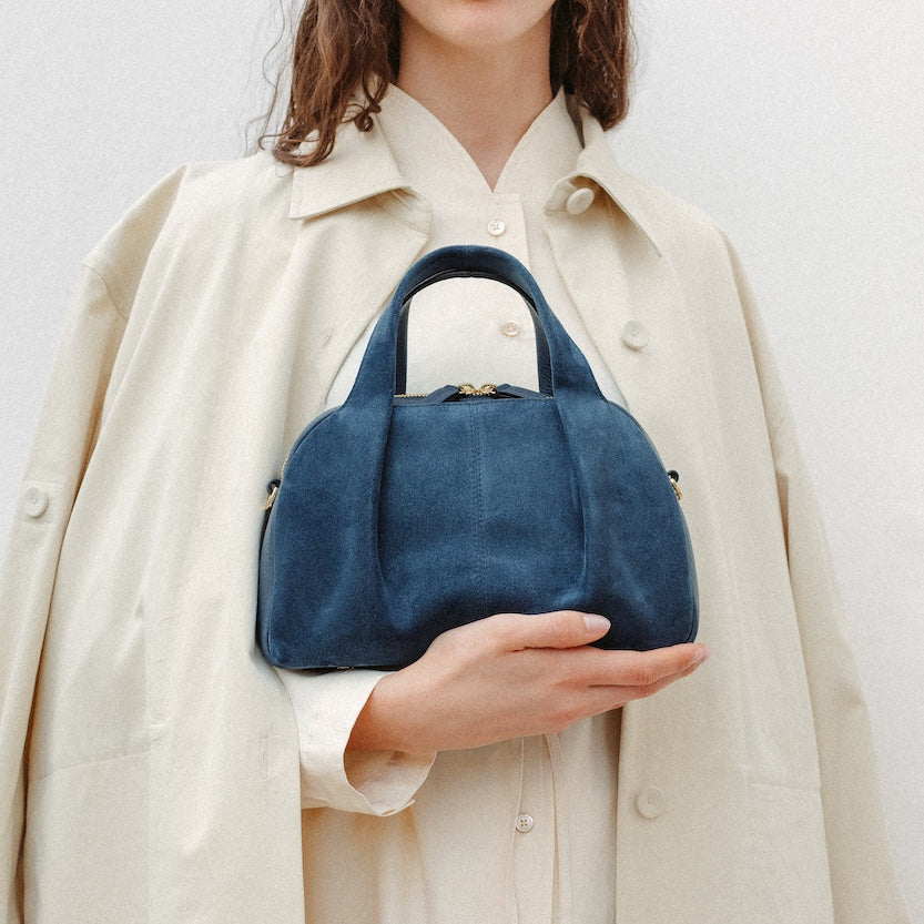 Model holding STOW London's Mini Bowling in Navy suede leather featuring a pleated front panel and gold-coated stainless steel hardware