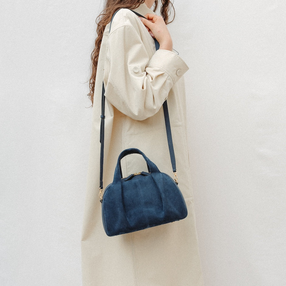 Model wearing STOW London's Mini Bowling in Navy suede leather on her shoulder featuring a pleated front panel and gold-coated stainless steel hardware