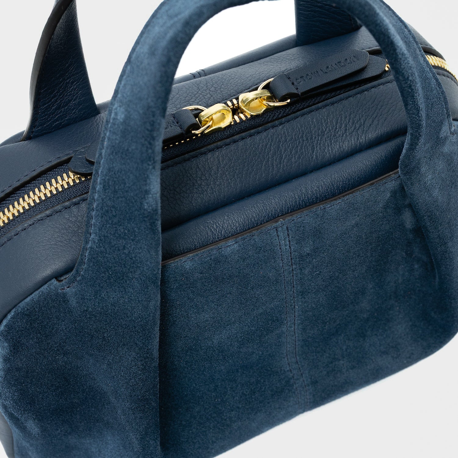 Close up of STOW London's Mini Bowling bag in Navy suede and smooth leather featuring the zip closure and STOW London embossed pull tabs with the external slip pocket in between the pleated panel on a light grey background