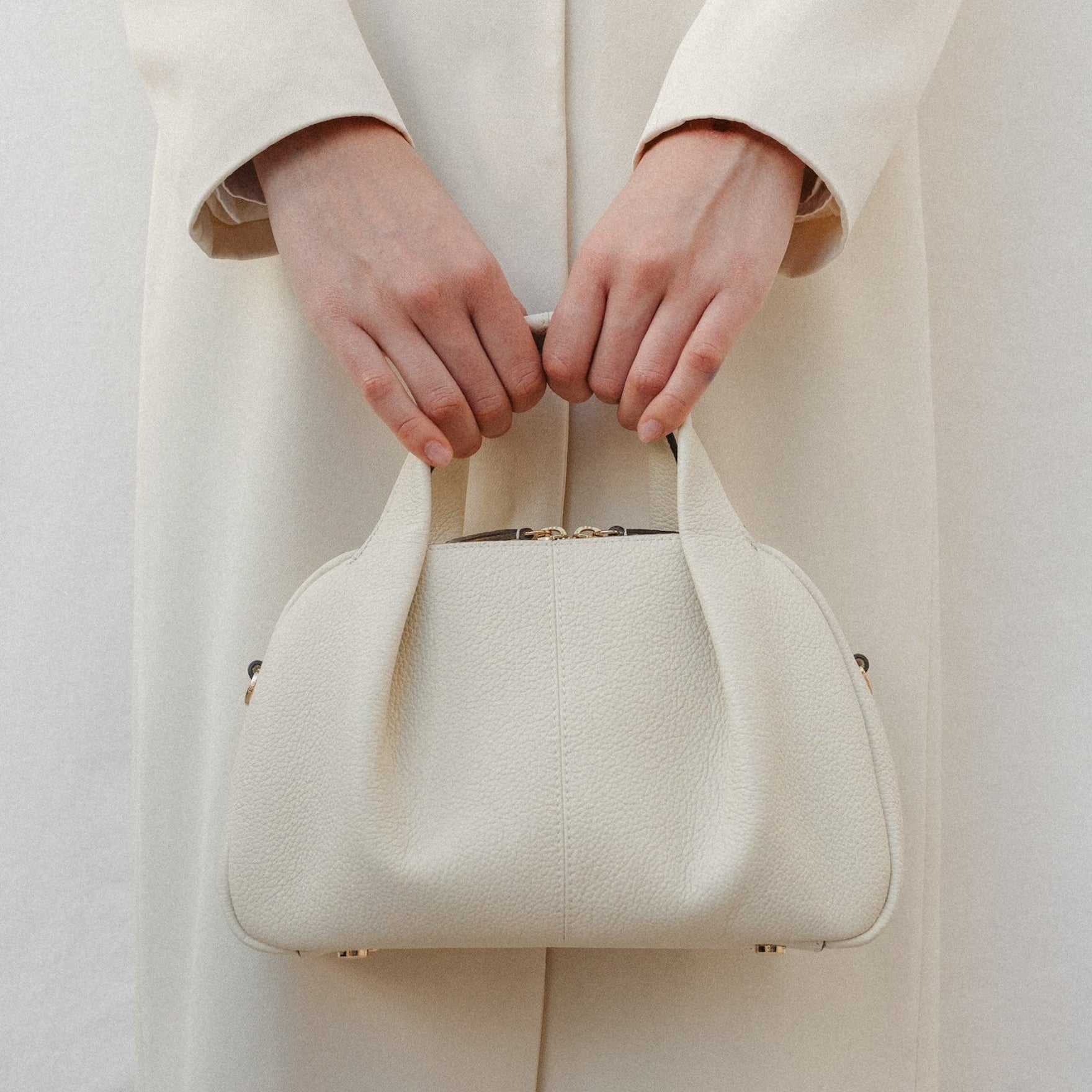 Model holding STOW London's Mini Bowling bag in Spring Moon pebbled leather by top handle featuring a pleated front panel and gold-coated stainless steel hardware