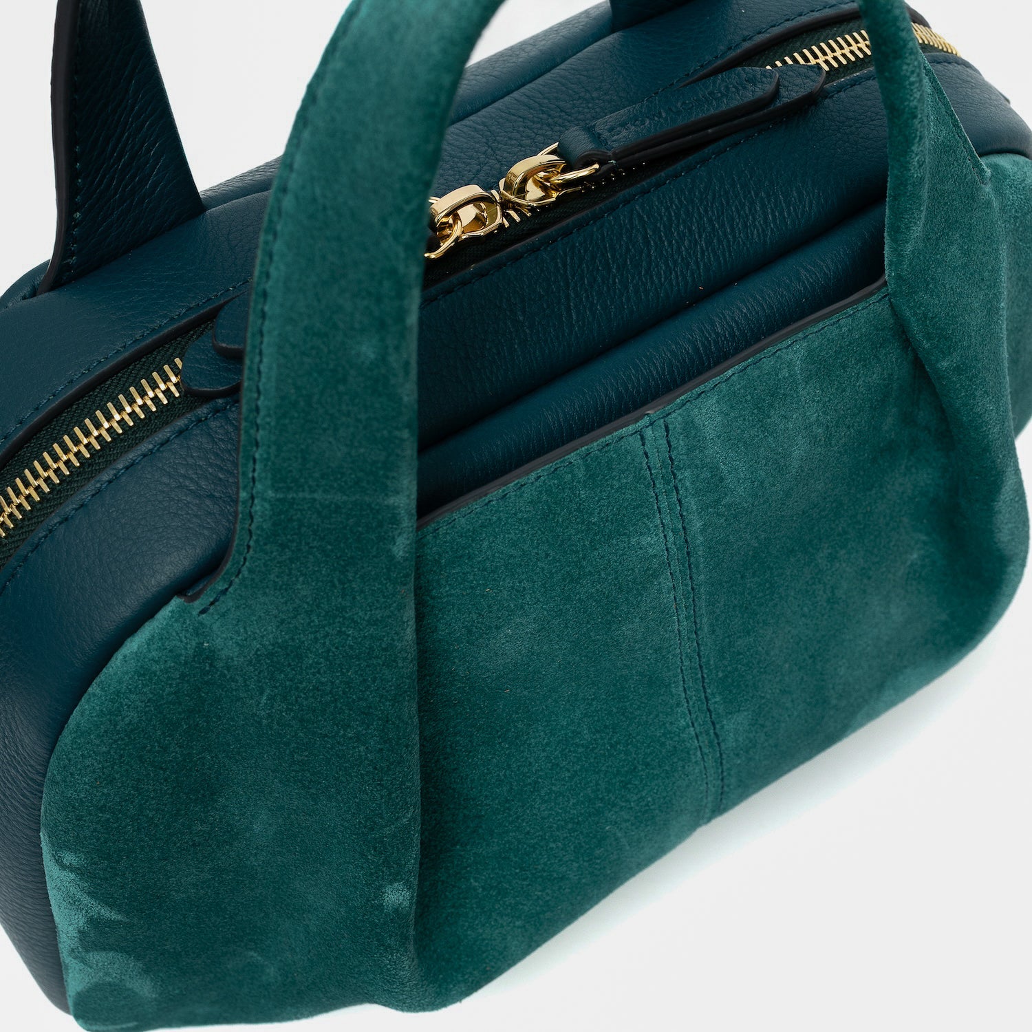 Close up of STOW London's Mini Bowling bag in Ocean Teal suede and smooth leather featuring a pleated front panel, the zip closure with STOW London embossed pull tabs on a light grey background