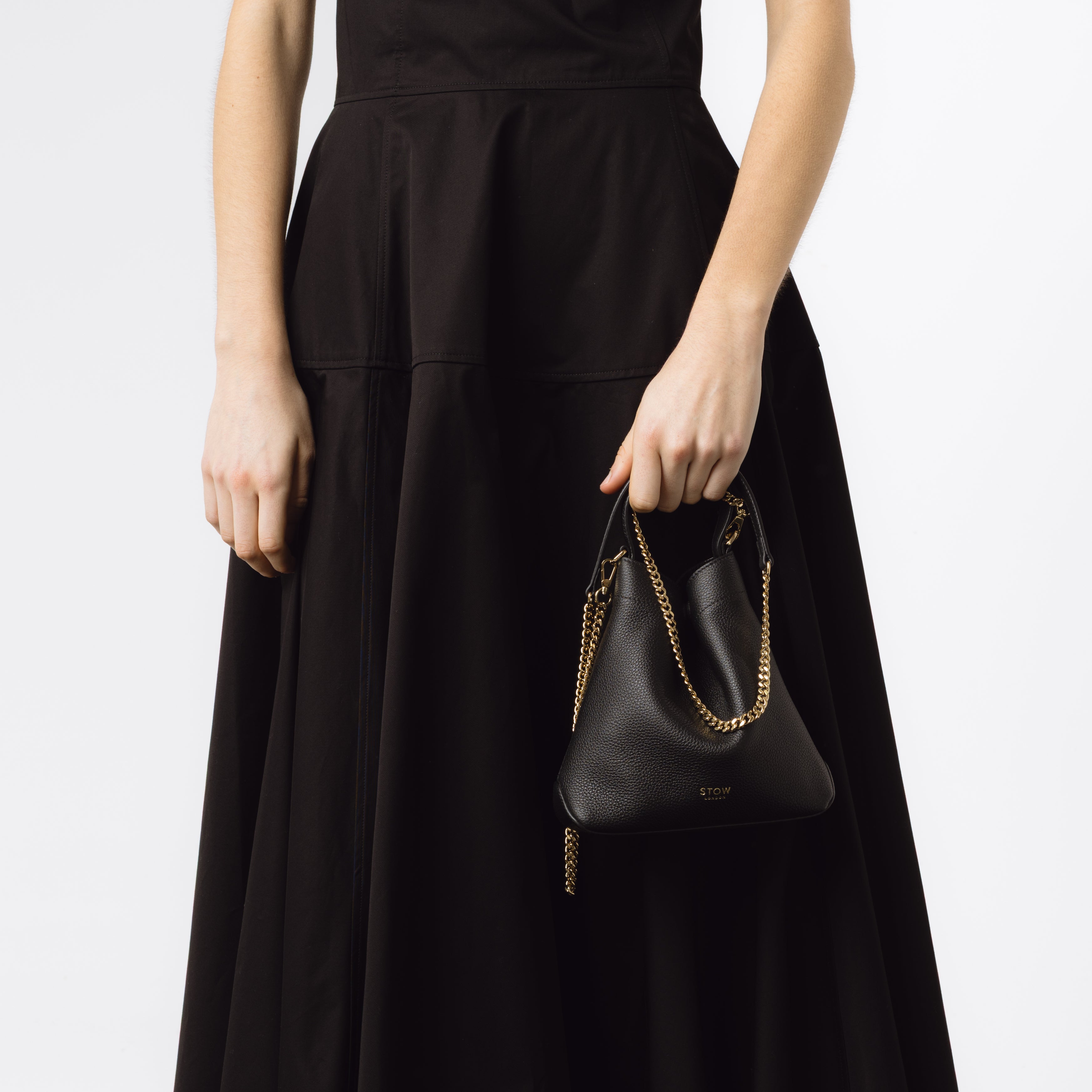 Model holding STOW London's Mini Curve in Black pebbled leather and detachable gold chain crossbody strap at her side on a white background