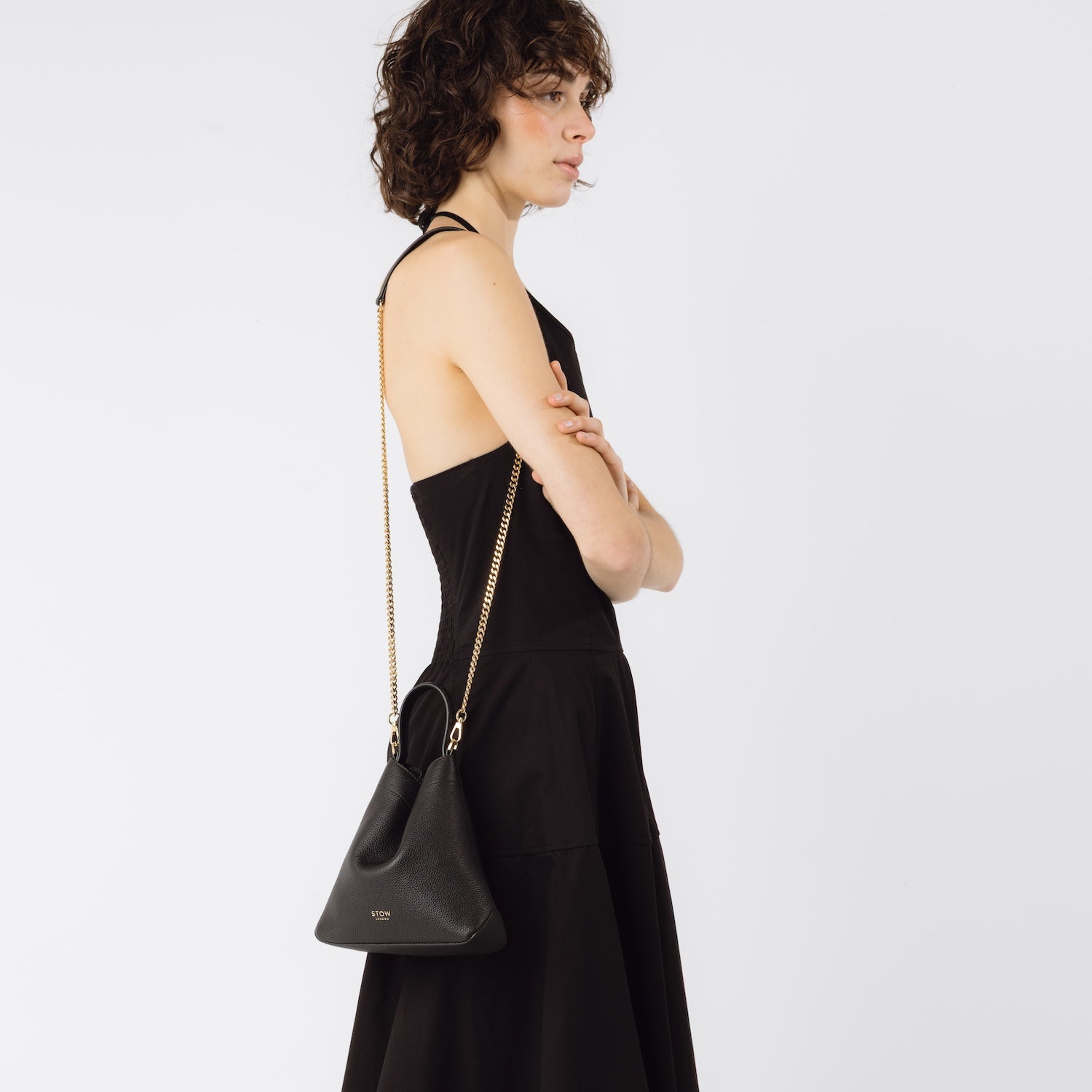 Model wearing STOW London's Mini Curve in Black pebbled leather on her shoulder by the detachable gold chain crossbody strap on a white background