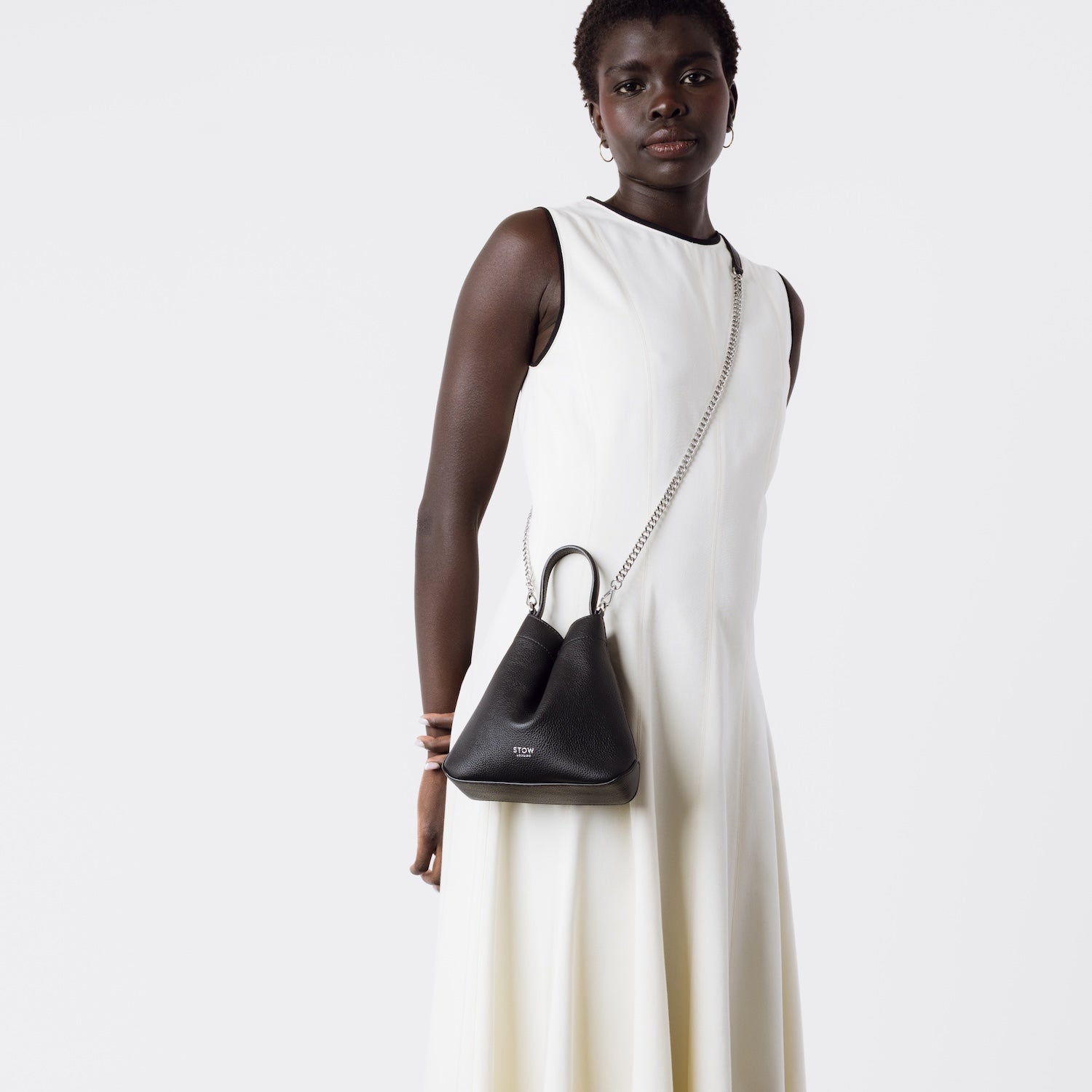 Model wearing Stow London's Mini Curve in Black pebbled leather crossbody by detachable silver chain crossbody strap on a white background