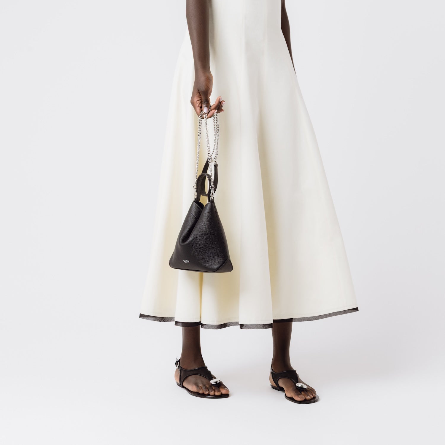 Model holding Stow London's Mini Curve in Black pebbled leather by the detachable silver chain crossbody strap on a white background