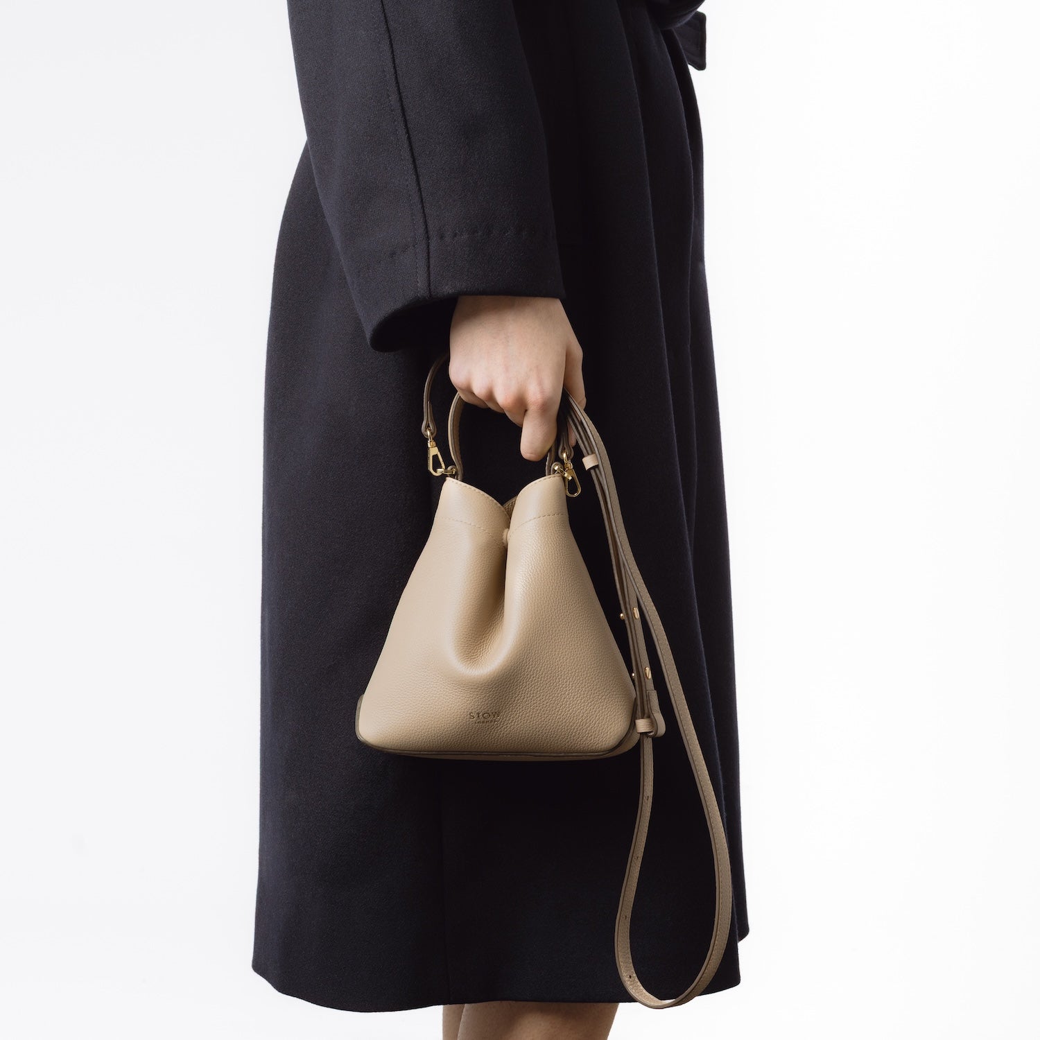 STOW London's Mini Curve and leather crossbody strap in Camel held by model's side by top handle, featuring gold-coated stainless steel hardware on a white background