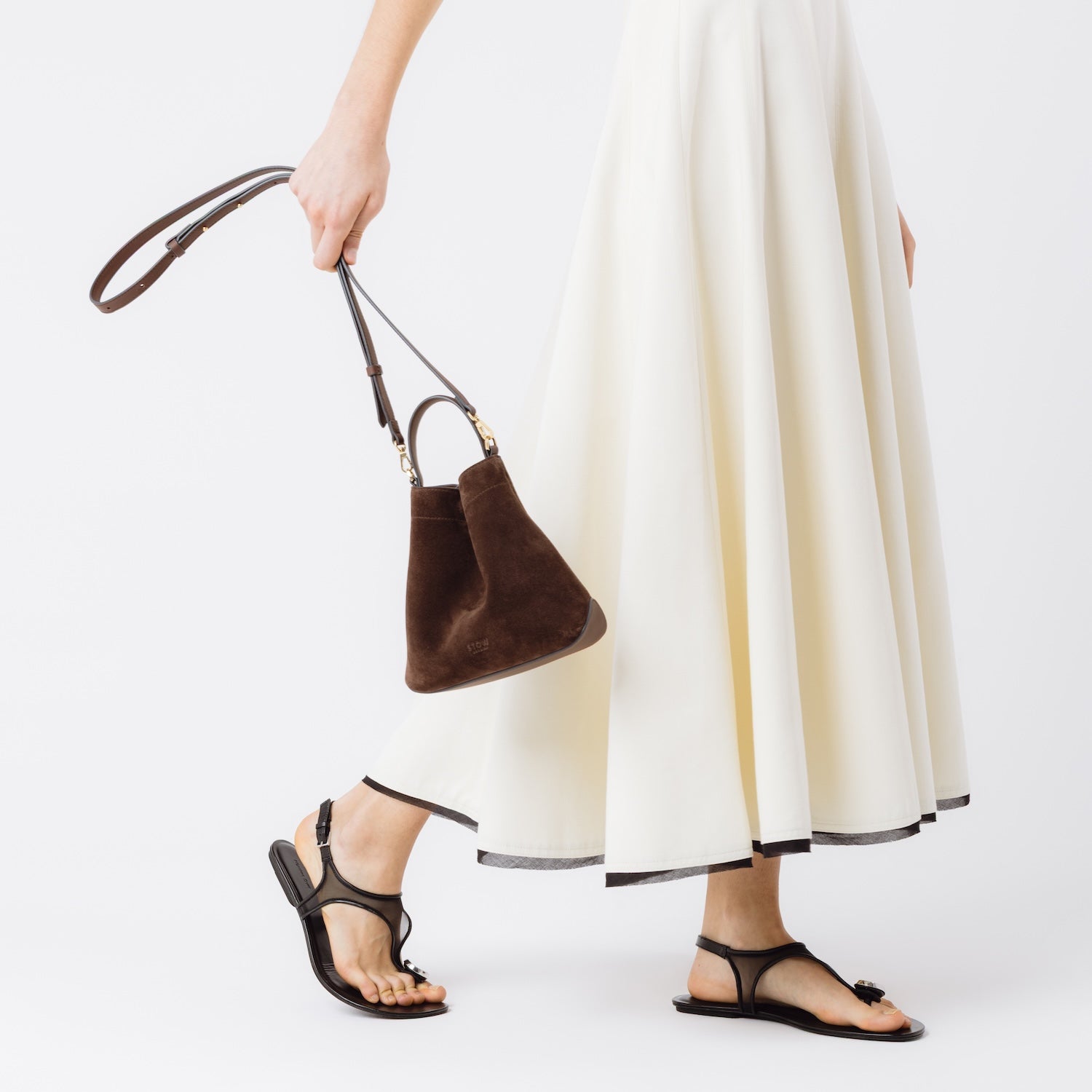 Model holding STOW London's Mini Curve in Chocolate suede leather by the detachable leather crossbody strap at her side on a white background