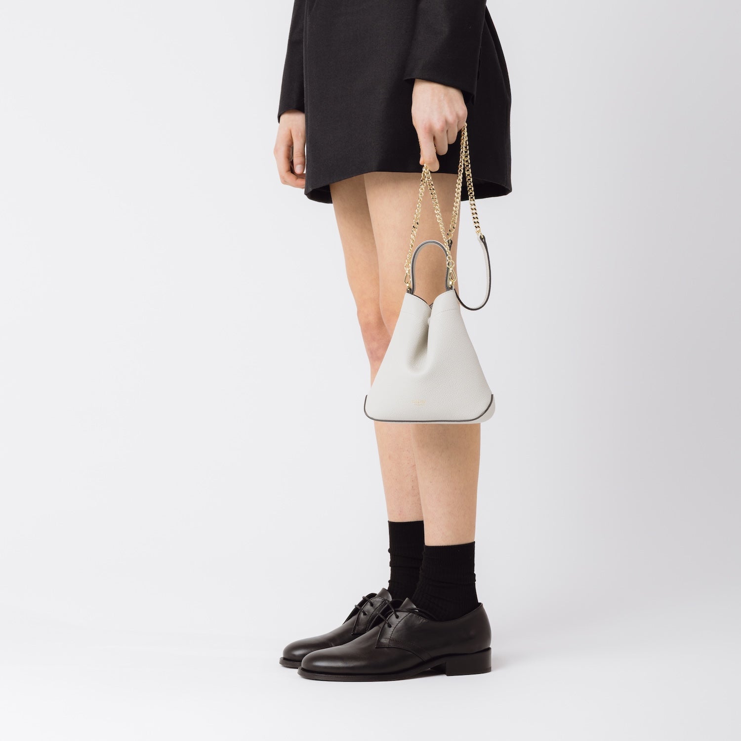 Model holding Stow London's Mini Curve in Cloud Grey pebbled leather by its detachable gold chain crossbody strap at her side on a white background