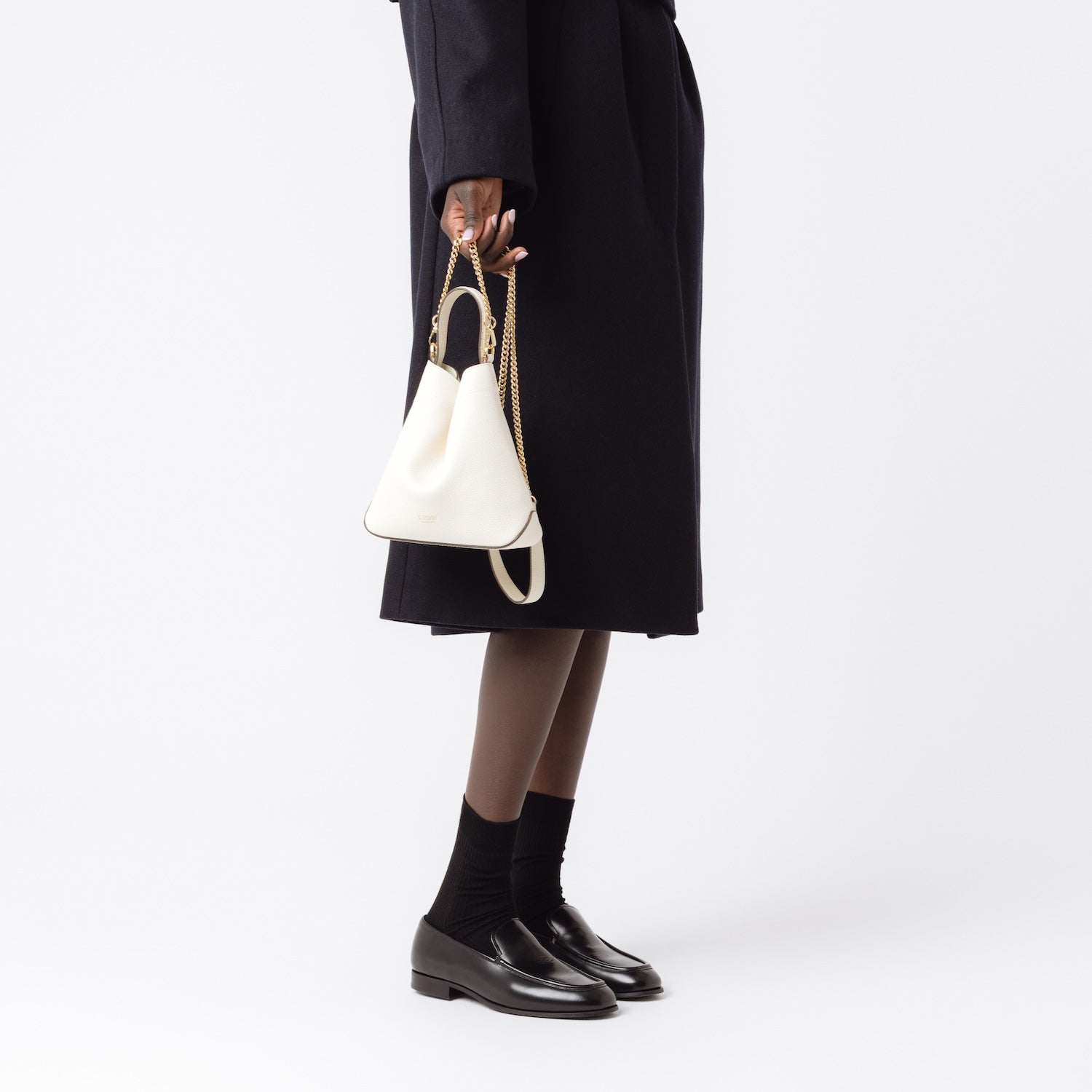 Model holding STOW London's Mini Curve in Spring Moon pebbled leather by the detachable gold chain crossbody strap at her side on a white background