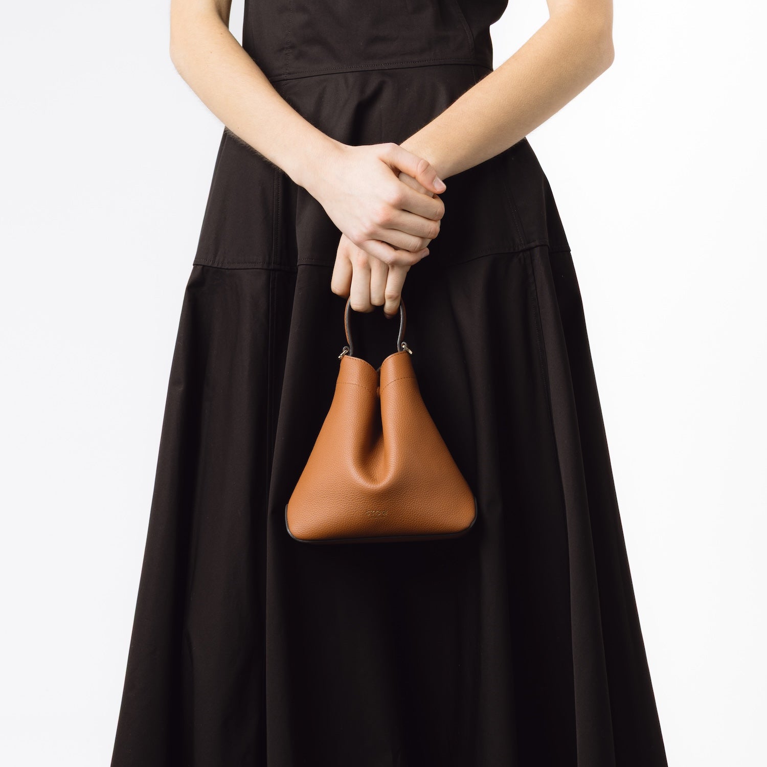 Model holding STOW London's Mini Curve bag in Earth Tan pebbled leather by its top handle on a white background