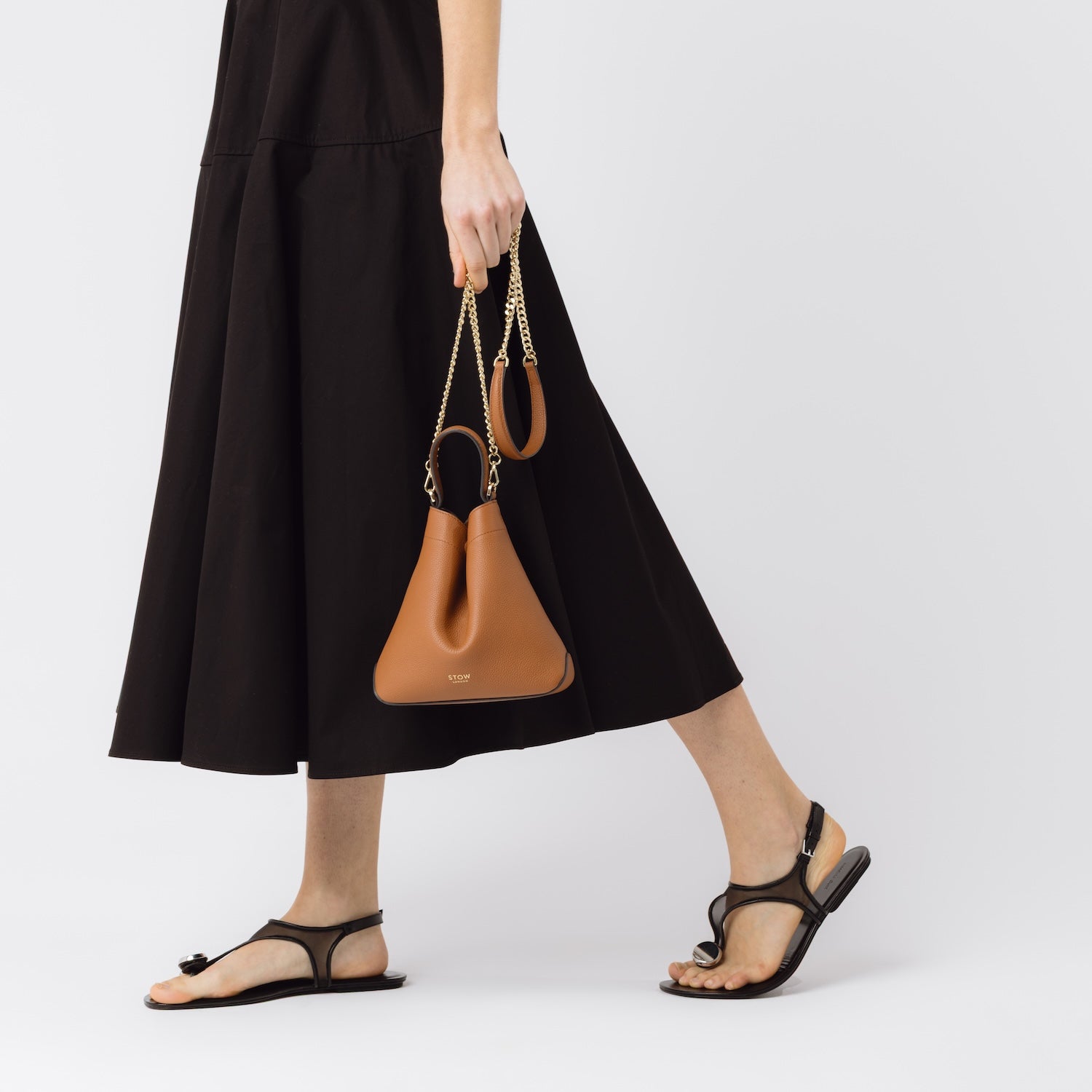 Model holding STOW London's Mini Curve bag in Earth Tan leather by its detachable gold-plated chain strap at her side on a white background