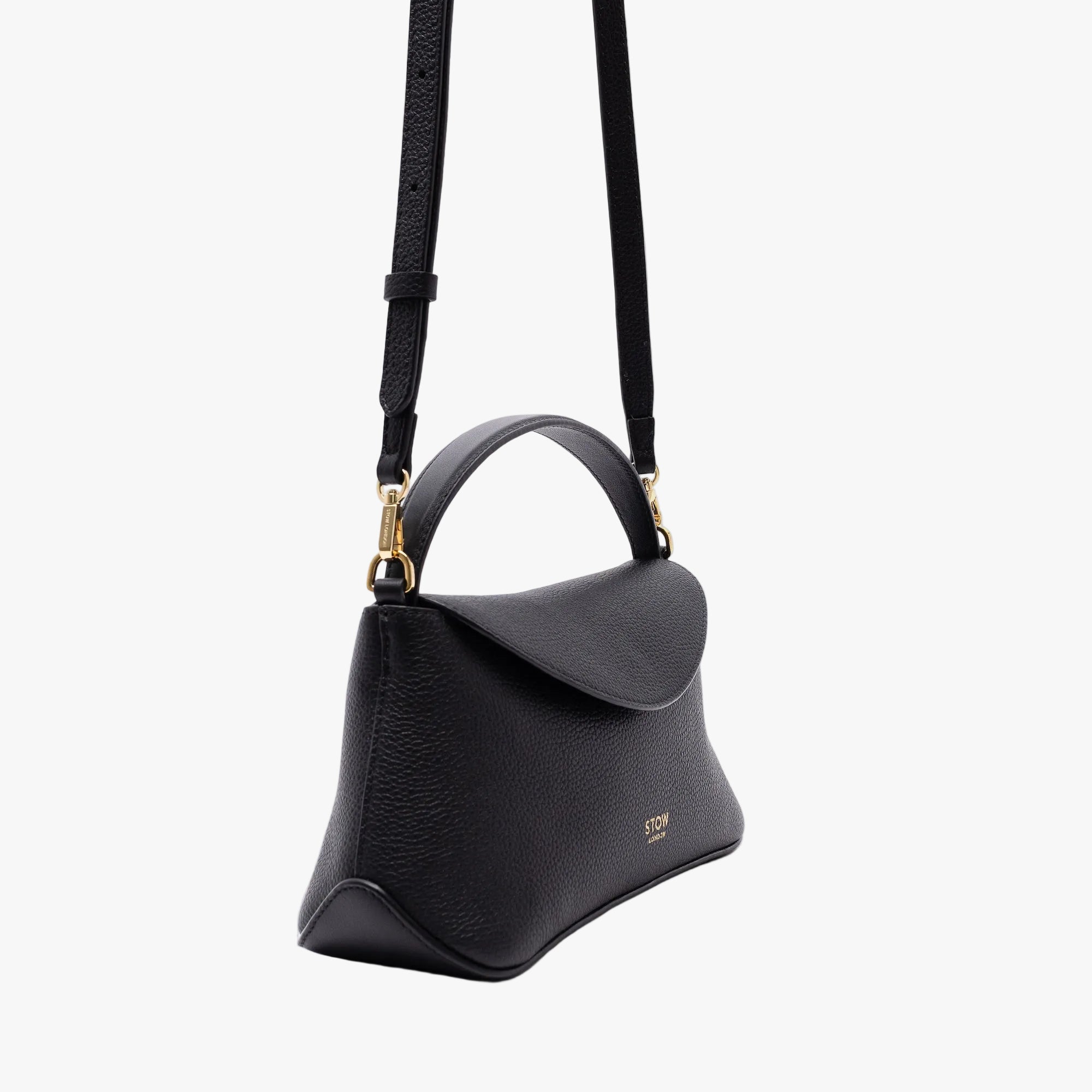 Off-axis view of STOW London's Mini East West in Black pebbled leather and detachable crossbody strap featuring gold-coated stainless steel hardware on a light grey background