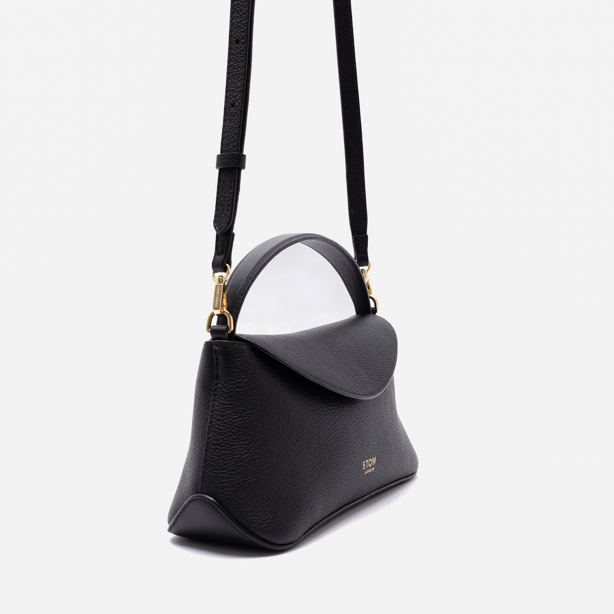 Off-axis view of STOW London's Mini East West in Black pebbled leather and detachable crossbody strap featuring gold-coated stainless steel hardware on a light grey background