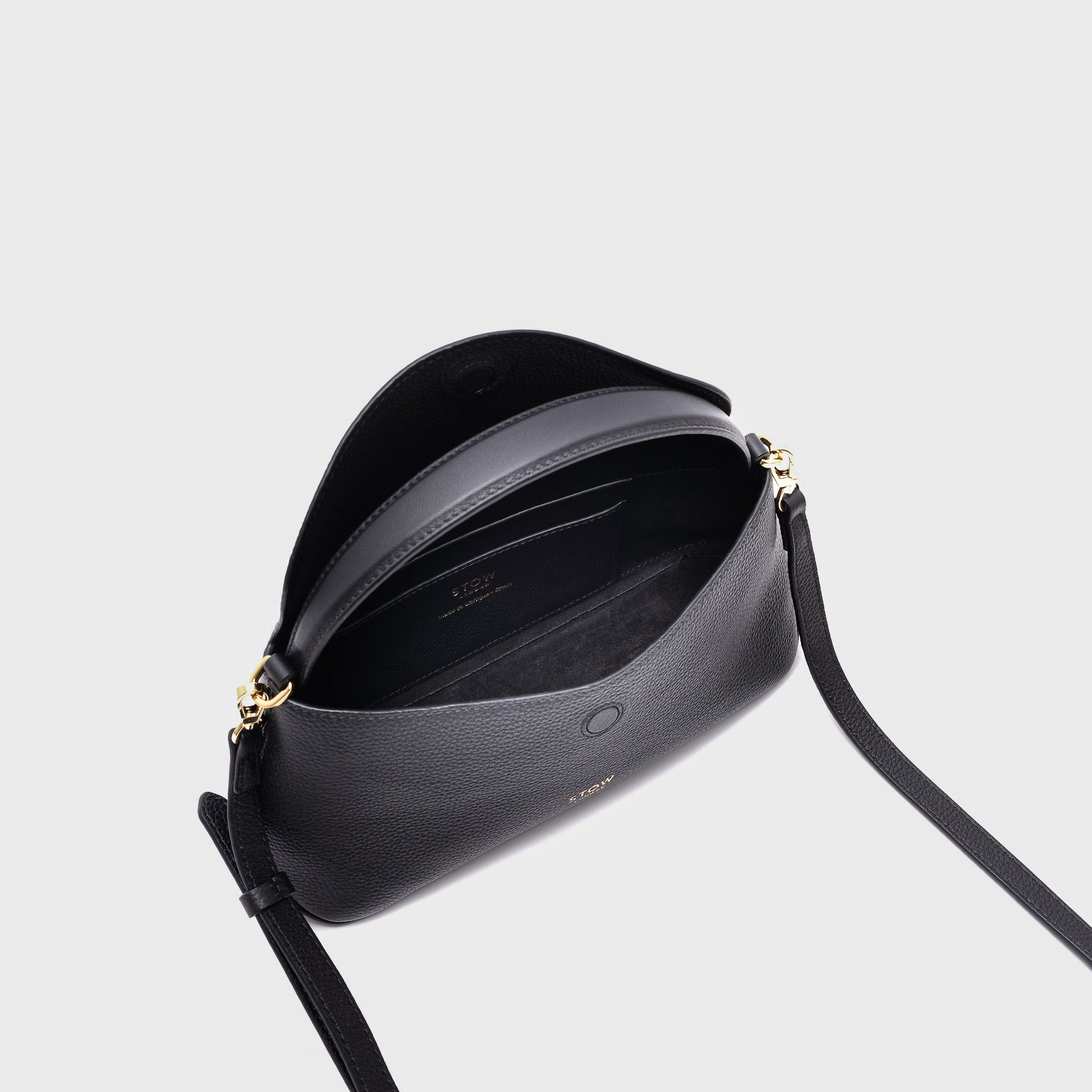 STOW London's Mini East West in Black pebbled leather with flap open showing suede interior and slip pocket featuring gold-coated stainless steel hardware on a light grey background
