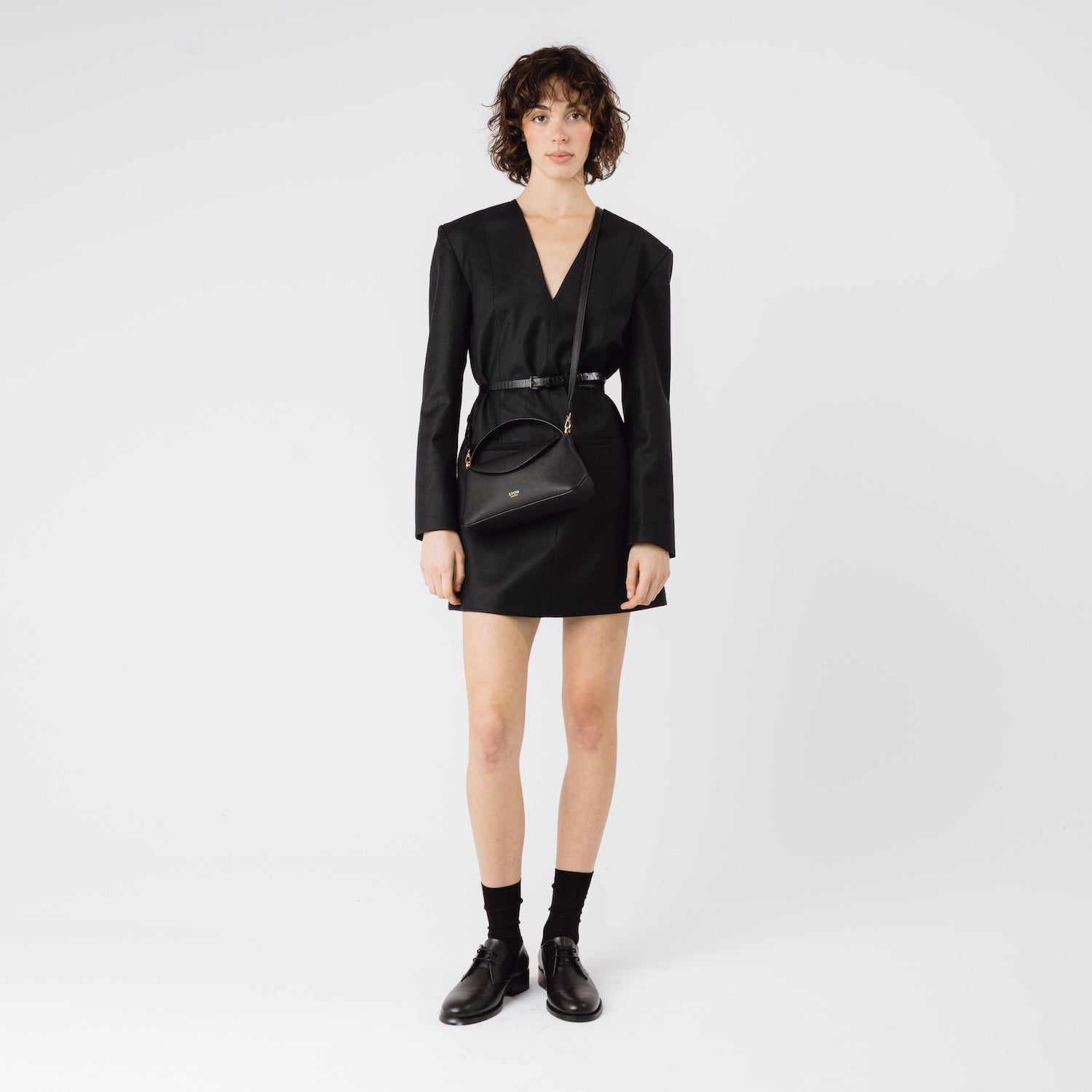 Model wearing STOW London's Mini East West in Black pebbled leather crossbody featuring gold-coated stainless steel hardware on a light grey background