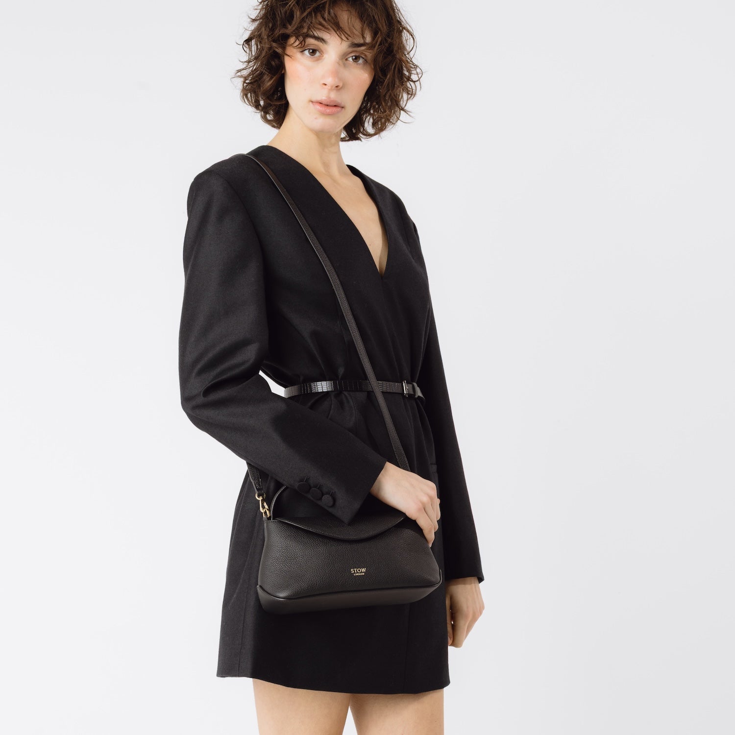 Model wearing STOW London's Mini East West in Black pebbled leather on her shoulder featuring gold-coated stainless steel hardware on a light grey background