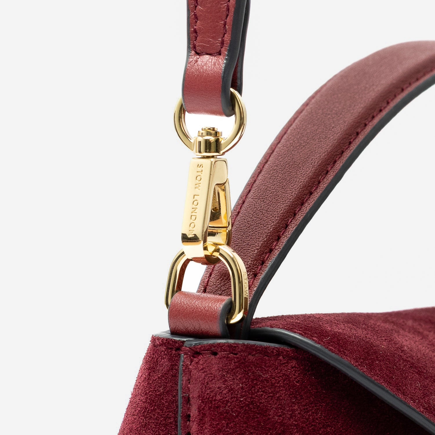 Close up of the detachable crossbody strap clip of STOW London's Mini East West in Burgundy suede leather featuring gold-coated stainless steel hardware on a light grey background