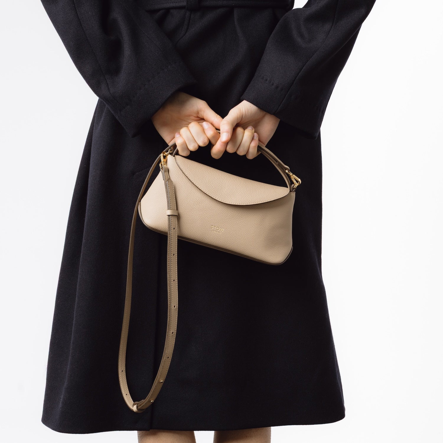 Model holding STOW London's Mini East West in Camel pebbled and smooth leather behind her back featuring the detachable crossbody strap and gold-coated stainless steel hardware on a white background