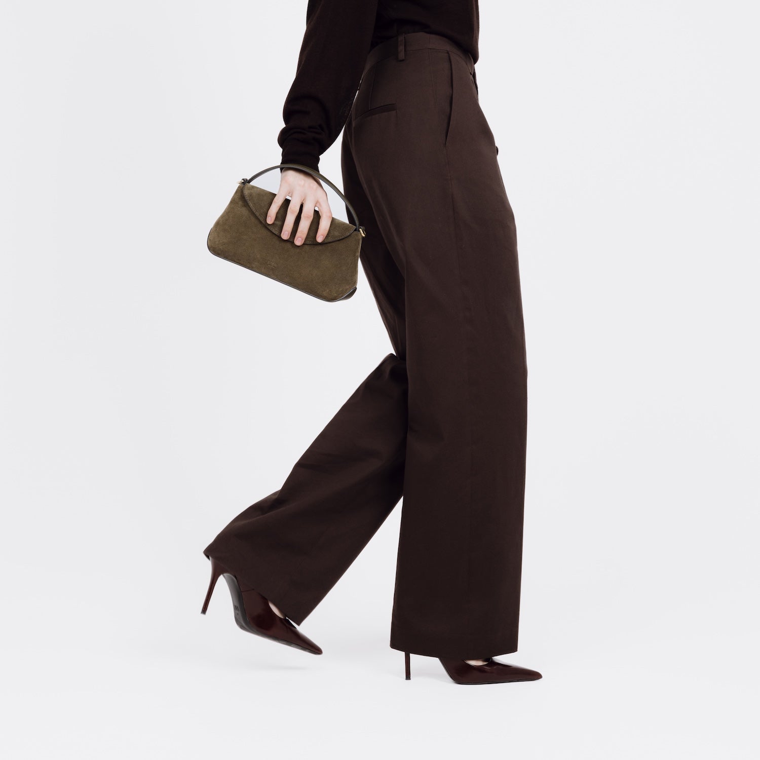 Model holding STOW London's Mini East West bag in Dark Green Suede leather at her side as a clutch featuring gold-coated stainless steel hardware on a white background