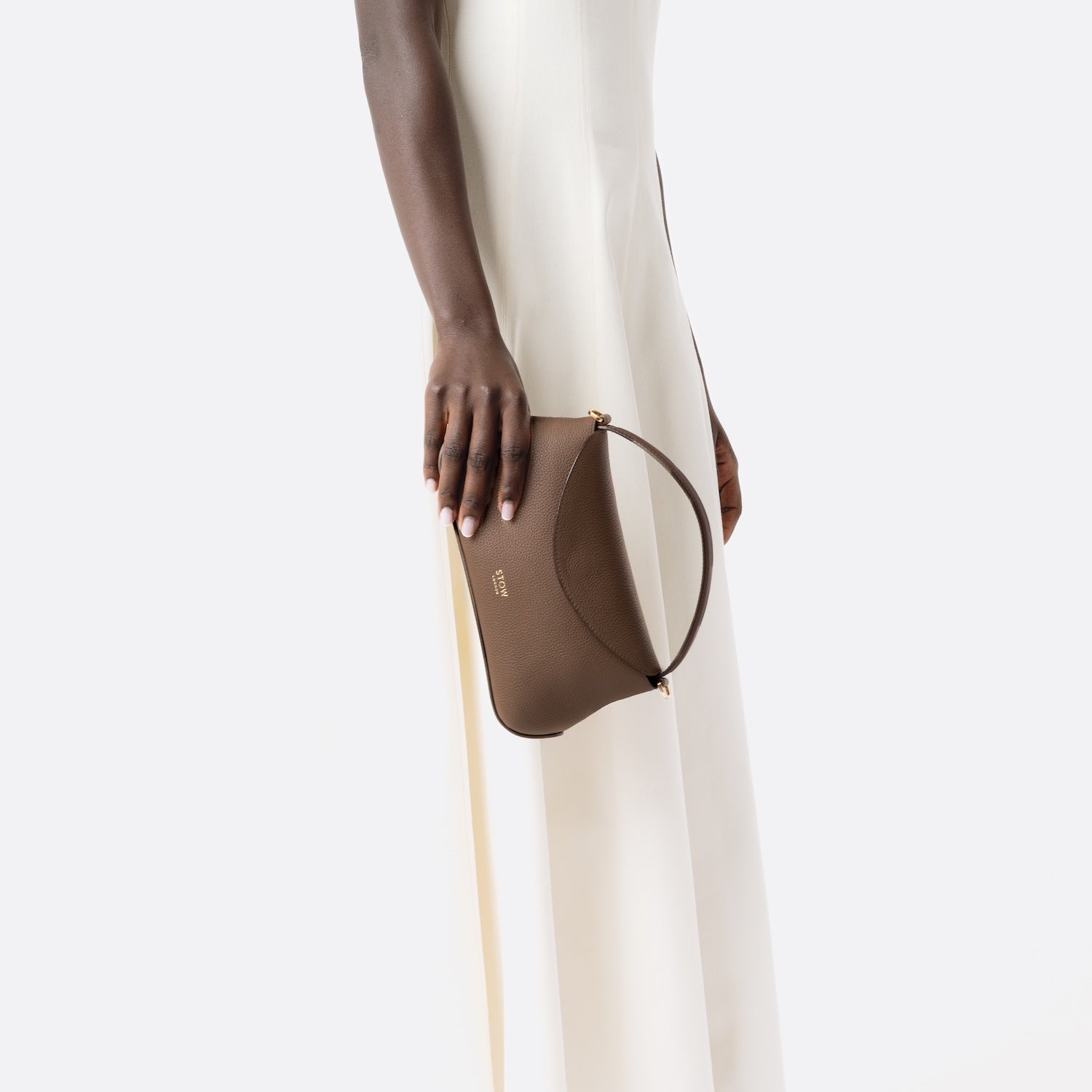 STOW London's Mini East West in Mocha pebbled and smooth leather held at model's side featuring gold-coated stainless steel hardware on a white background