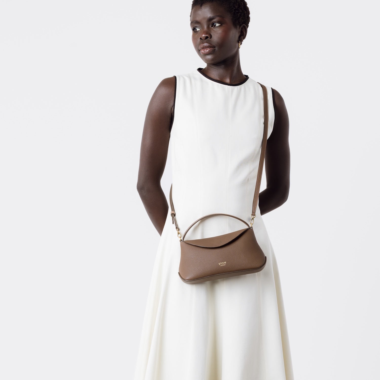 STOW London's Mini East West in Mocha pebbled and smooth leather worn crossbody by model featuring gold-coated stainless steel hardware on a white background