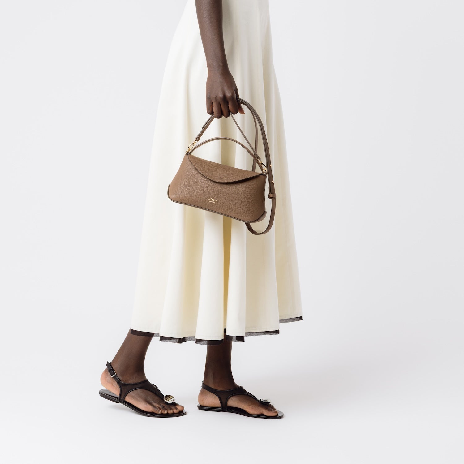 STOW London's Mini East West in Mocha pebbled and smooth leather held by detachable crossbody strap at model's side featuring gold-coated stainless steel hardware on a white background