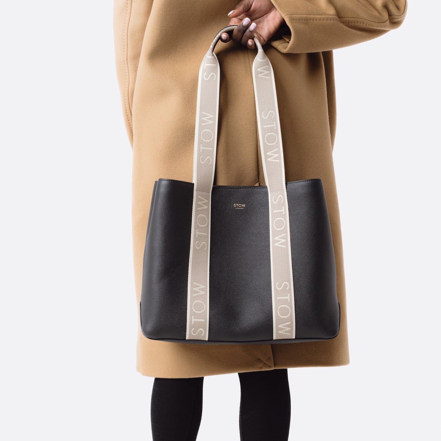Model holding STOW London's Nastro Tote bag in Black pebbled leather with beige nastro handles featuring 'STOW' branding on a white background