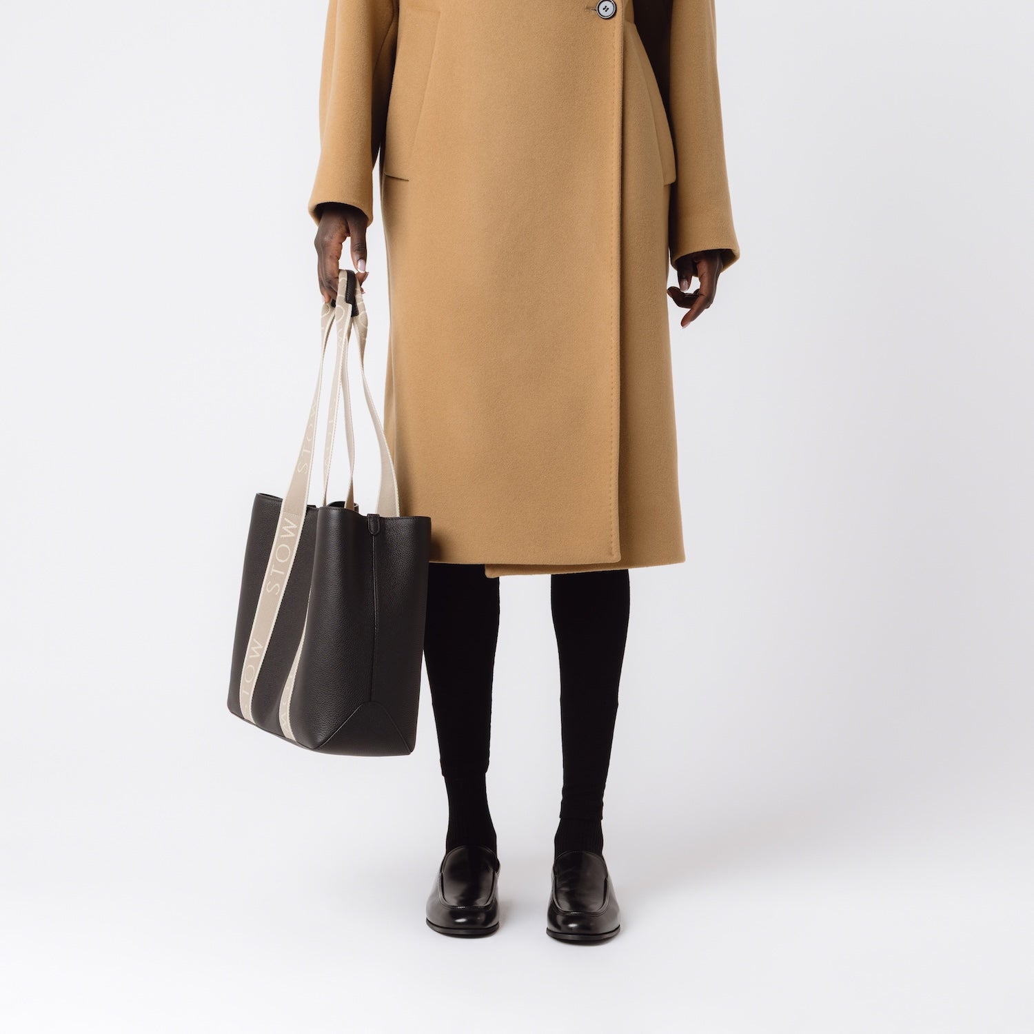 Model holding STOW London's Nastro Tote bag in Black pebbled leather at her side with beige nastro handles featuring 'STOW' branding on a white background