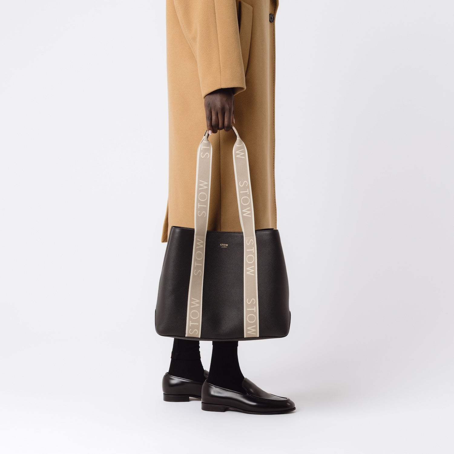 Model holding STOW London's Nastro Tote bag in Black pebbled leather with beige nastro handles featuring 'STOW' branding on a white background