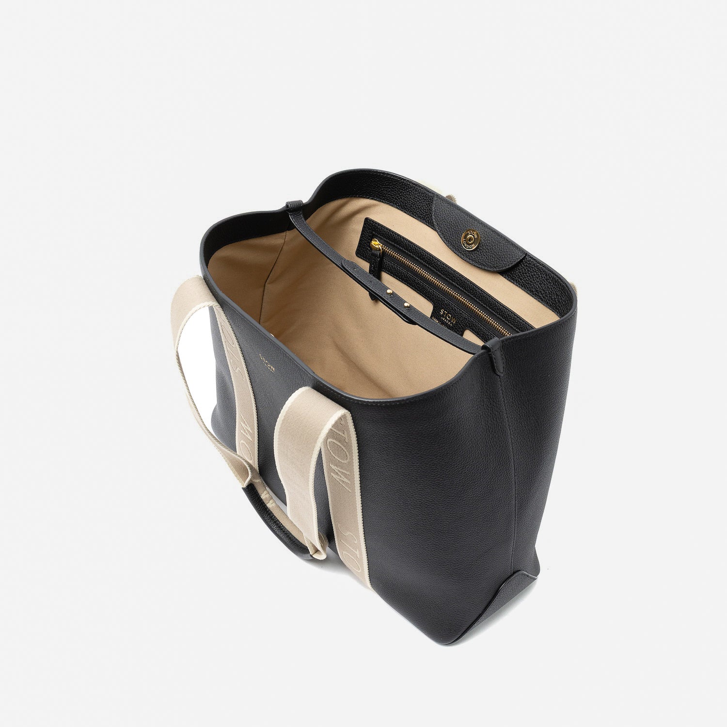 Top angled view of STOW London's Nastro Tote bag in Black pebbled leather with magnets open showing light beige cotton interior and zip pocket on a light grey background