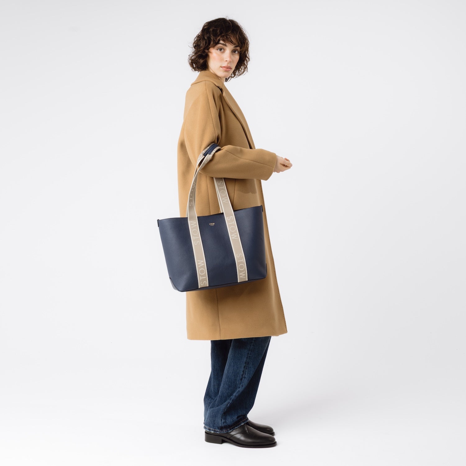 Model wearing STOW London's Nastro Tote bag in Navy pebbled leather with beige nastro handles featuring 'STOW' branding on her arm on a white background