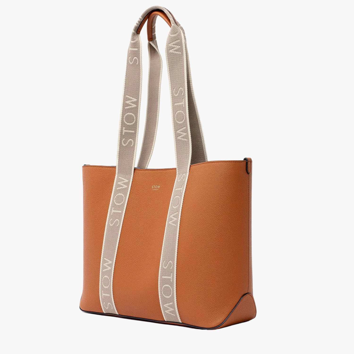 Off-axis view of STOW London's Nastro Tote bag in Earth Tan pebbled leather with beige nastro handles featuring 'STOW' branding on a light grey background