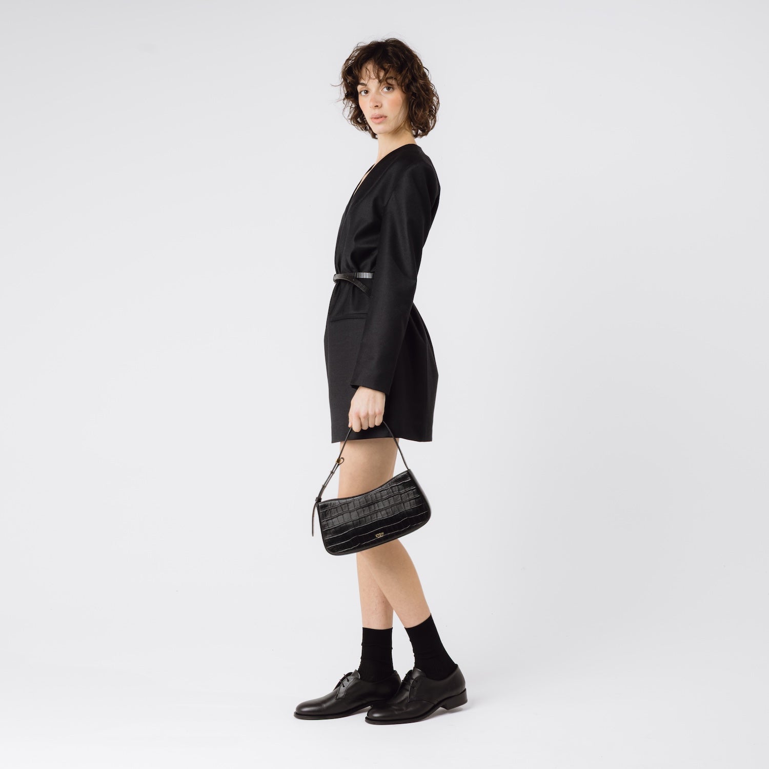 Model holding STOW London's Post Shoulder bag in Black Embossed Croc leather at her side featuring gold-coated stainless steel hardware on a white background
