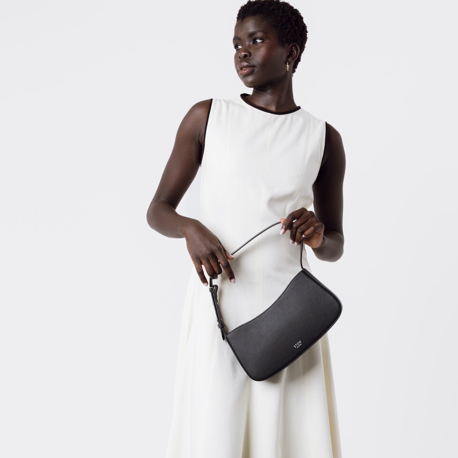 Model holding STOW London's Post Shoulder bag in Black smooth leather at her front featuring silver hardware on a white background