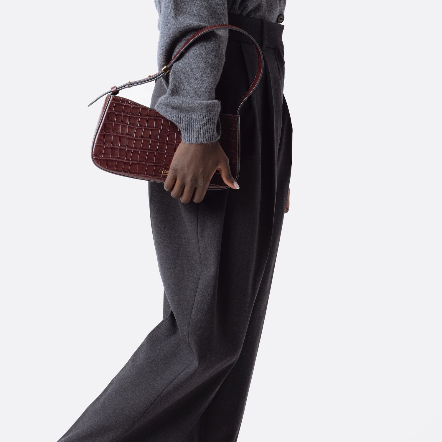 Model holding STOW London's Post Shoulder bag in Burgundy Embossed Croc leather in her hand at her side featuring gold-coated stainless steel hardware on a white background