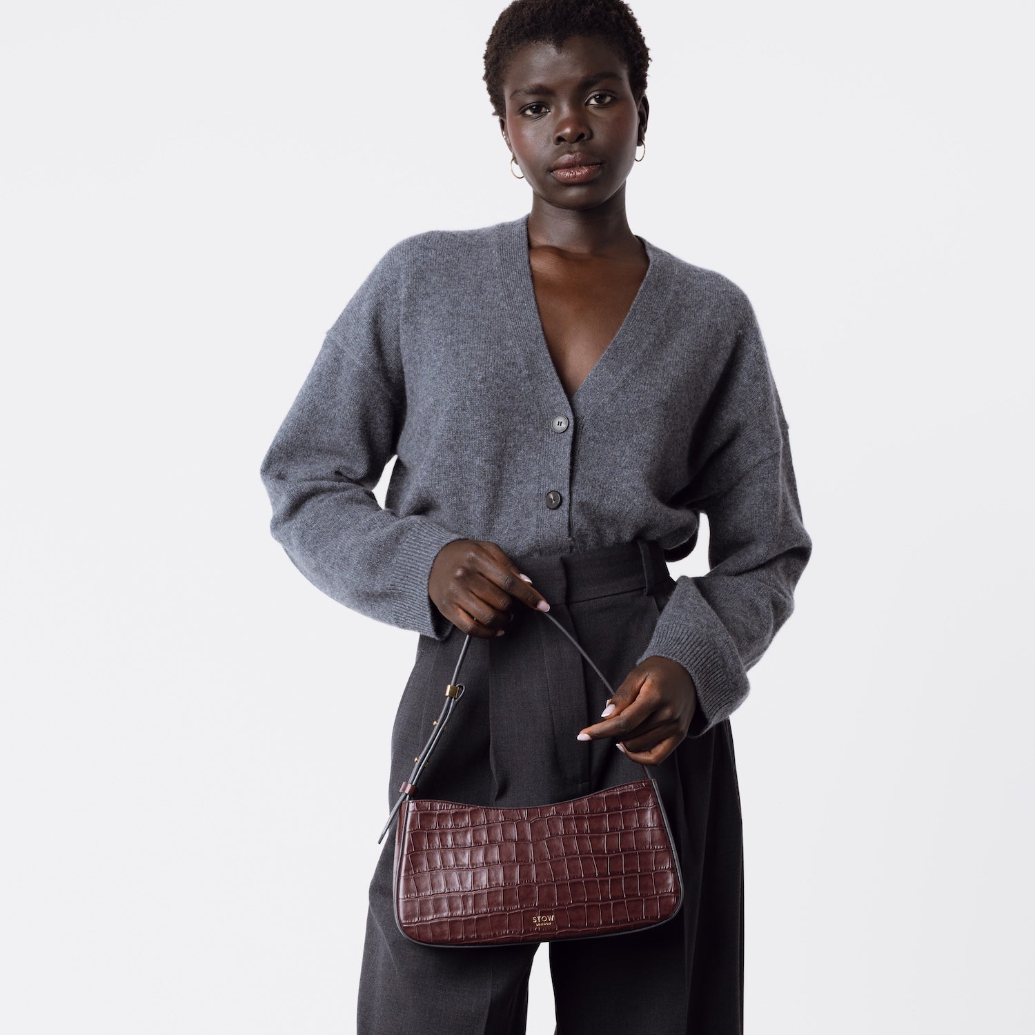 Model holding STOW London's Post Shoulder bag in Burgundy Embossed Croc leather at her front featuring gold-coated stainless steel hardware on a white background