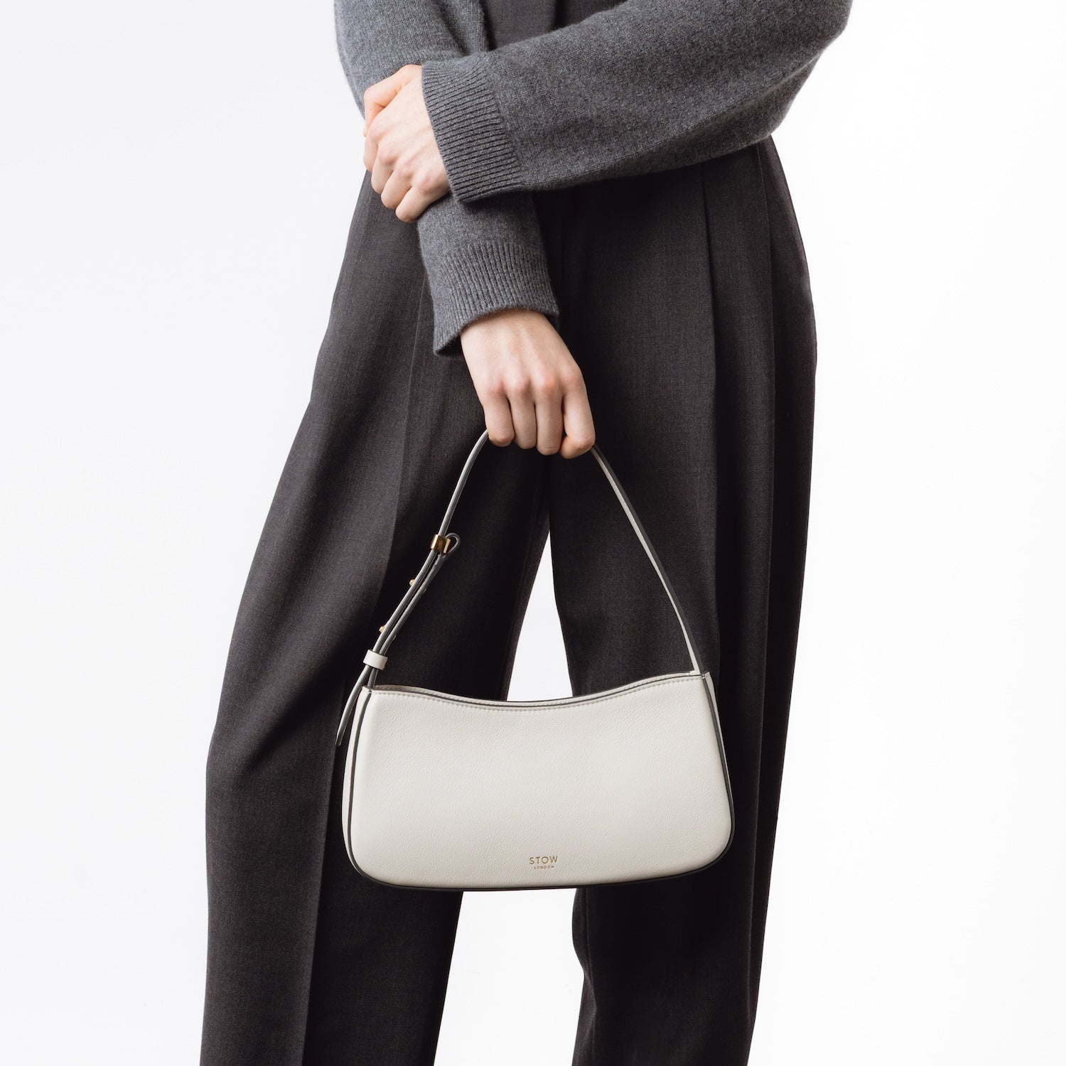 Model posing with STOW London's Post Shoulder bag in Cloud Grey smooth leather at her front featuring gold-coated stainless steel hardware on a white background
