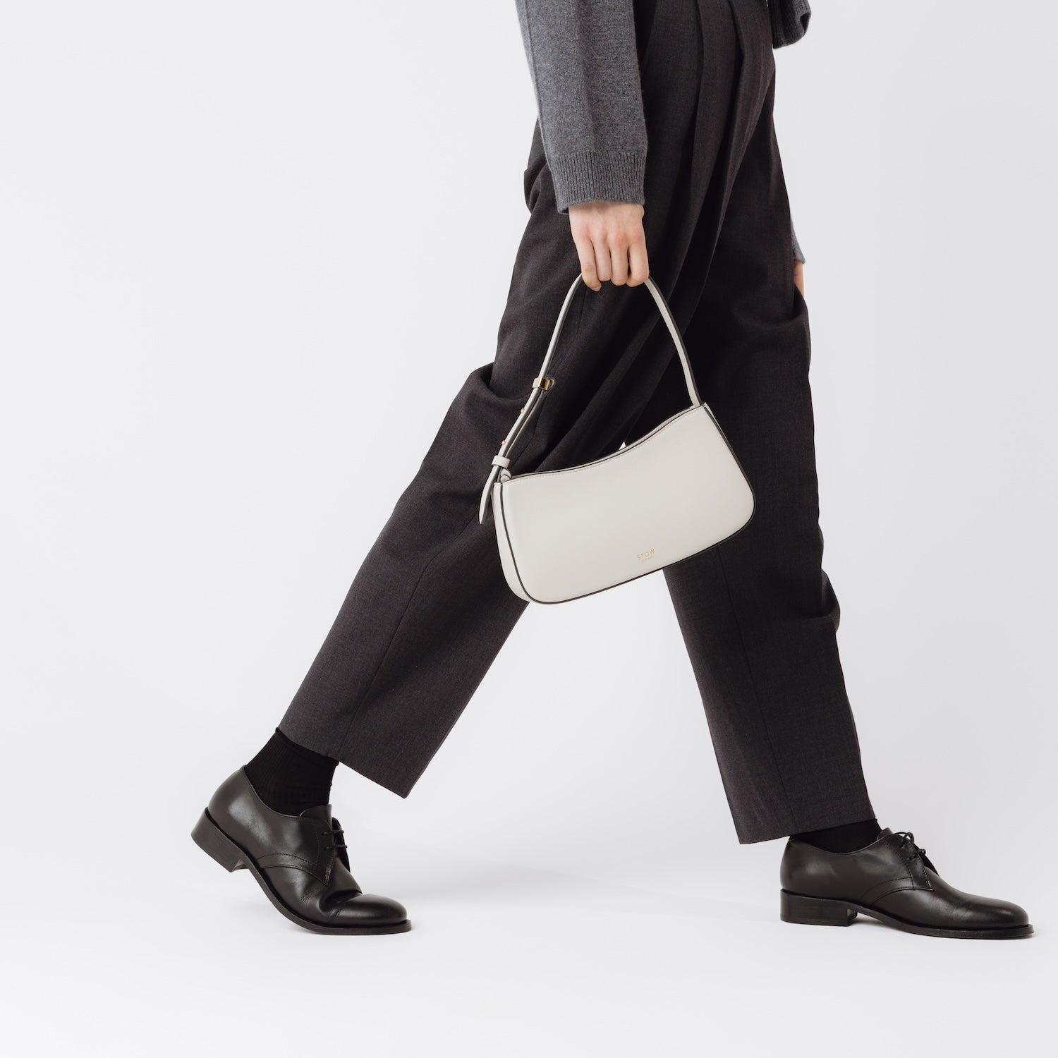 Model holding STOW London's Post Shoulder bag in Cloud Grey smooth leather at her side featuring gold-coated stainless steel hardware on a white background