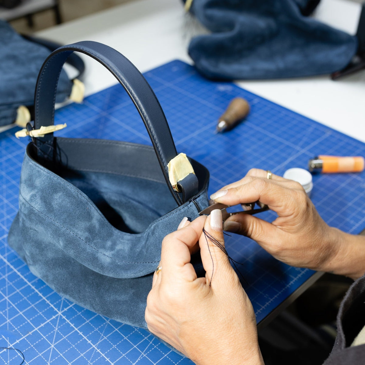 A close-up of a person's hands crafting a dark blue suede handbag on a blue cutting mat, with leatherworking tools visible in the background.