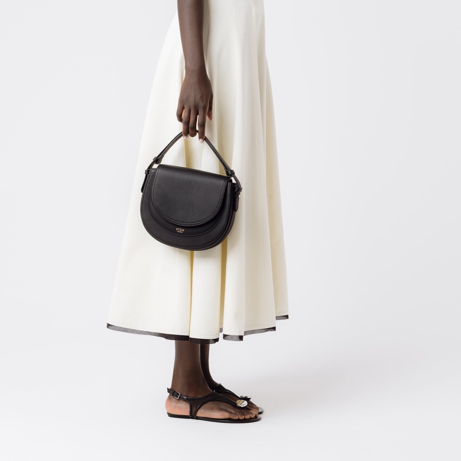 Model holding STOW London's Saddle bag in Black smooth leather by the detachable top handle at her side featuring gold-coated stainless steel hardware on a white background
