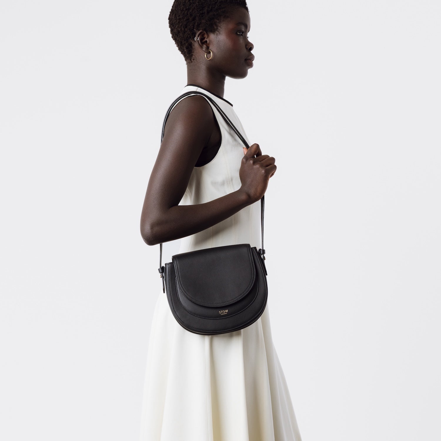 Model wearing STOW London's Saddle bag in Black smooth leather on her shoulder by the detachable and adjustable crossbody strap featuring gold-coated stainless steel hardware on a white background