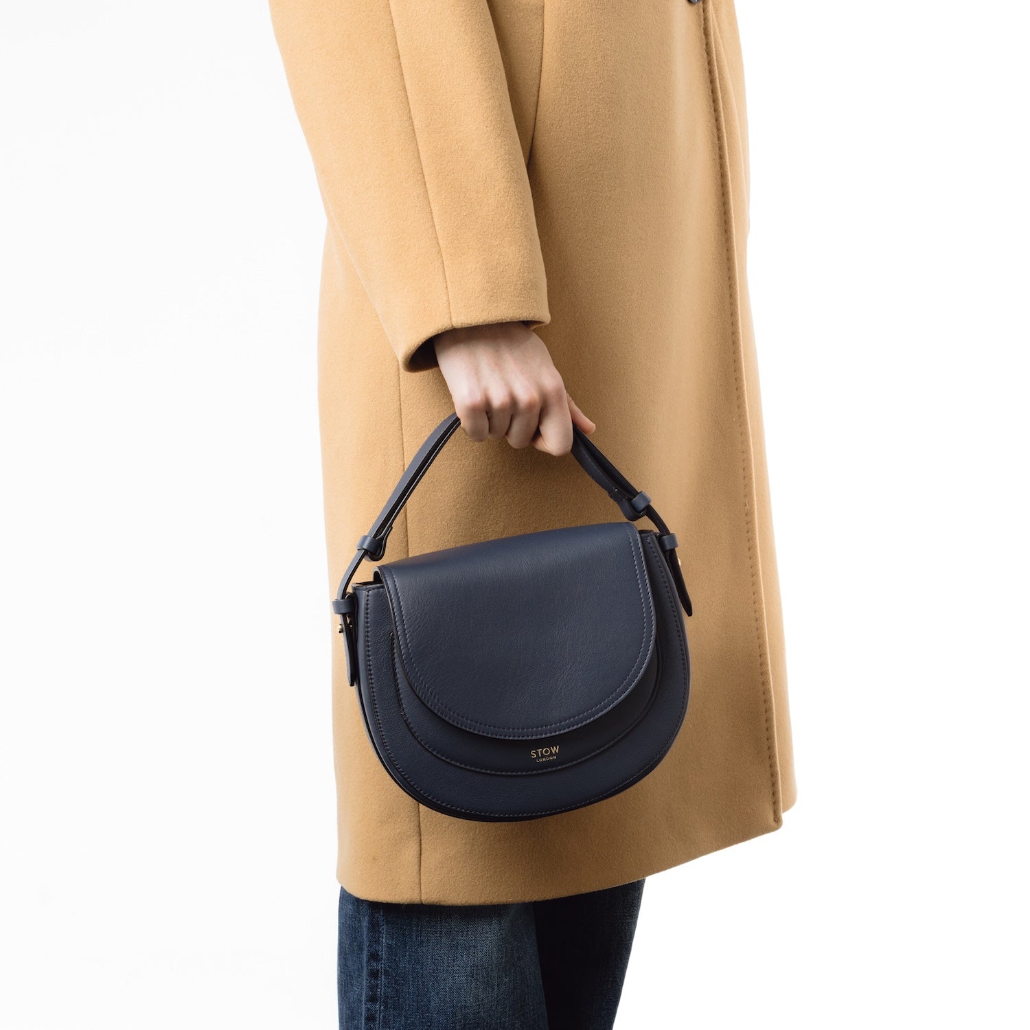 Model holding STOW London's Saddle bag in Navy smooth leather by the detachable top handle at her side featuring gold-coated stainless steel hardware on a white background