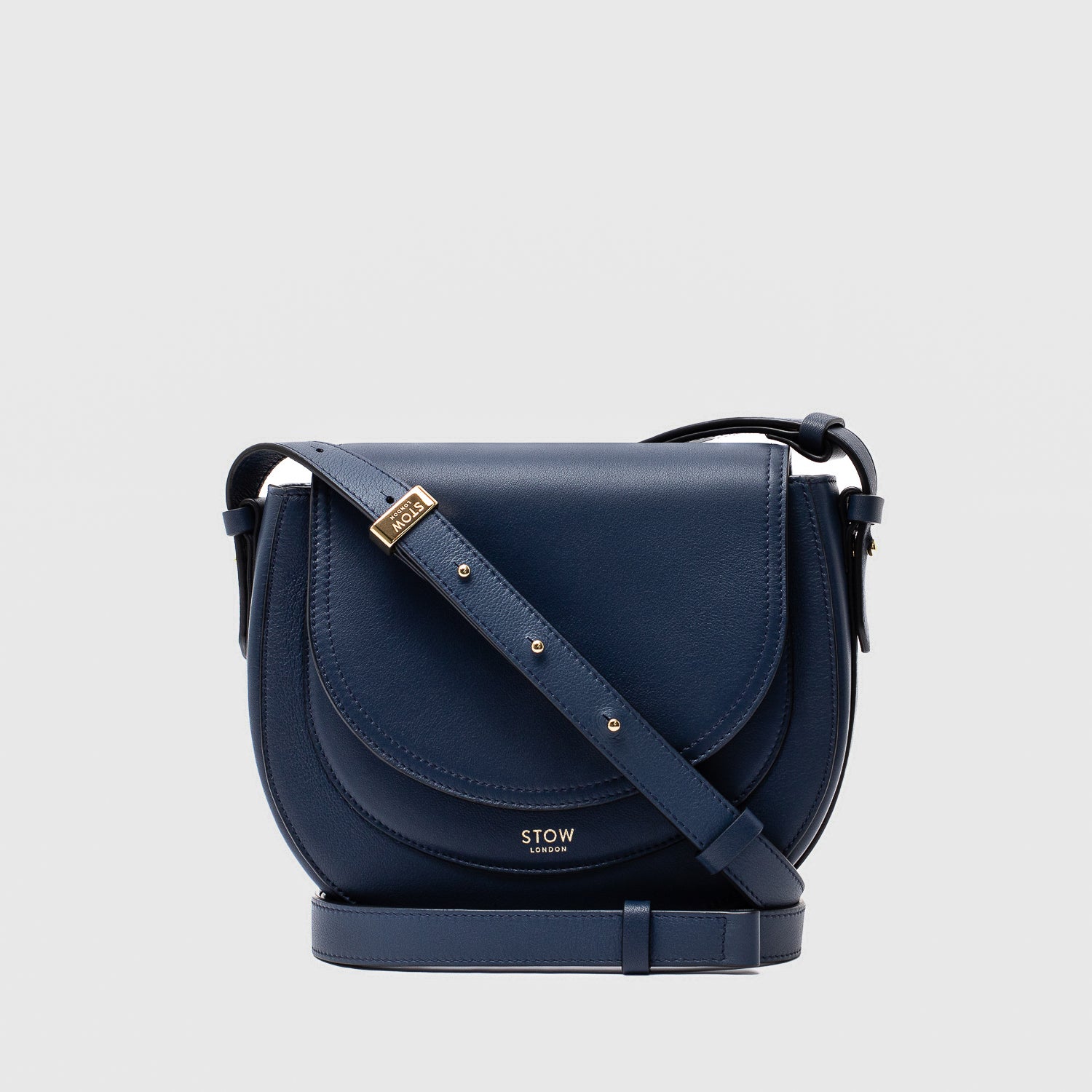 STOW London's Saddle bag in Navy smooth leather and detachable crossbody strap featuring gold-coated stainless steel hardware on a light grey background