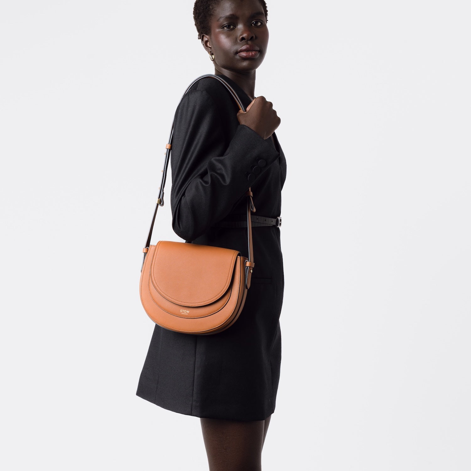 Model wearing STOW London's Saddle bag in Earth Tan smooth leather on her shoulder with its detachable crossbody strap on a white background